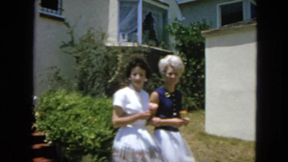 In 1963 San Francisco home-movie footage, two women stand in a sunny residential yard near shrubs and a light-colored house with a small cat.