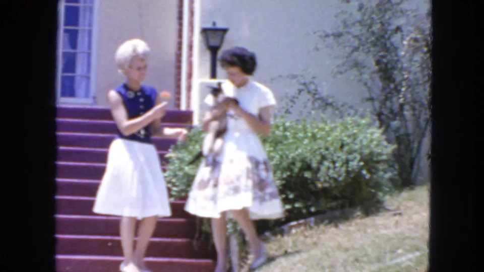 Two women in skirts stand beside purple-painted steps in San Francisco, 1963, with one holding a small cat near the house entrance.