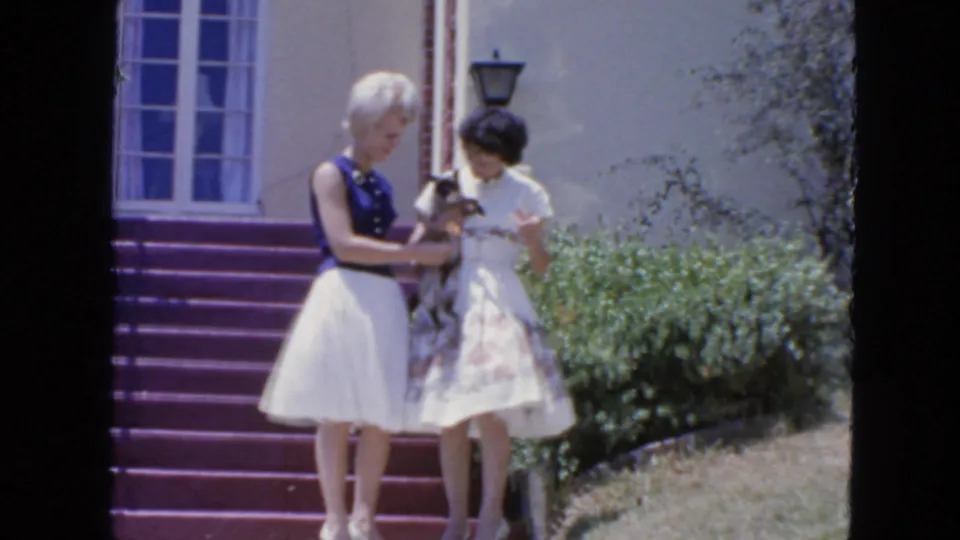 A 1963 San Francisco home-movie frame shows two women beside purple steps, focused on a small cat between them.