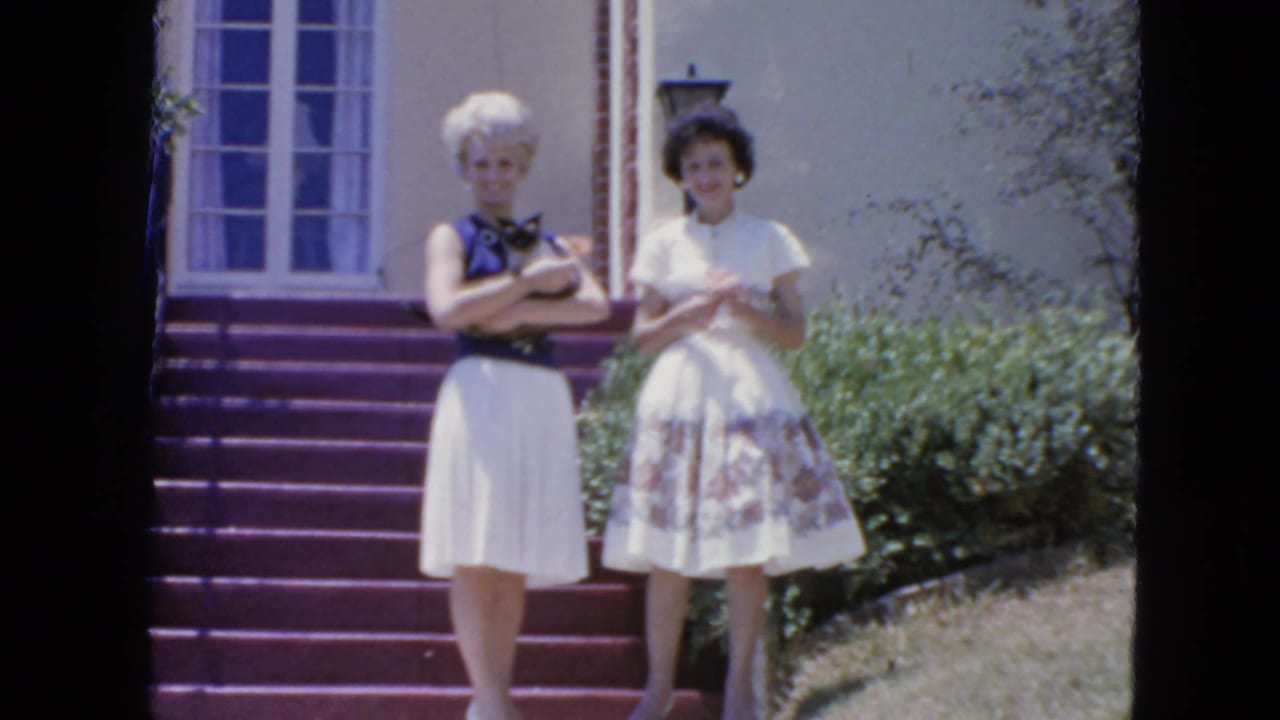 Two adult women in 1960s dresses stand on purple steps outside a San Francisco home, one holding a Siamese cat.