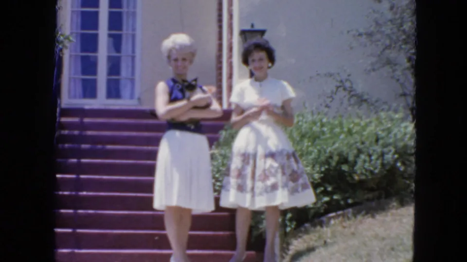 Two adult women in 1960s dresses stand on purple steps outside a San Francisco home, one holding a Siamese cat.