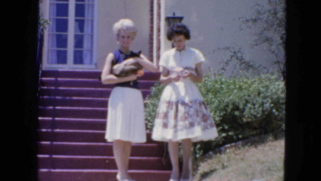 On purple exterior steps in 1963 San Francisco, a blonde woman holds a Siamese cat as her companion looks down at her hands.