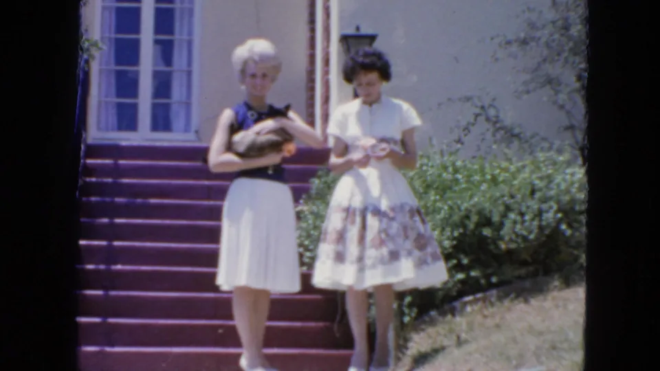 On purple exterior steps in 1963 San Francisco, a blonde woman holds a Siamese cat as her companion looks down at her hands.
