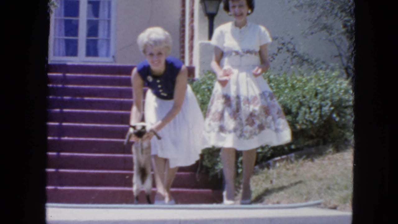 Two women stand near red-purple steps as one bends toward a small cat.