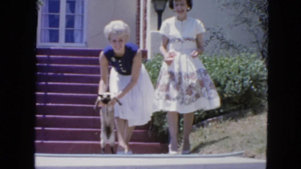 1963 San Francisco home-movie frame showing two women outside a house, with one bending to hold a small cat near porch steps.