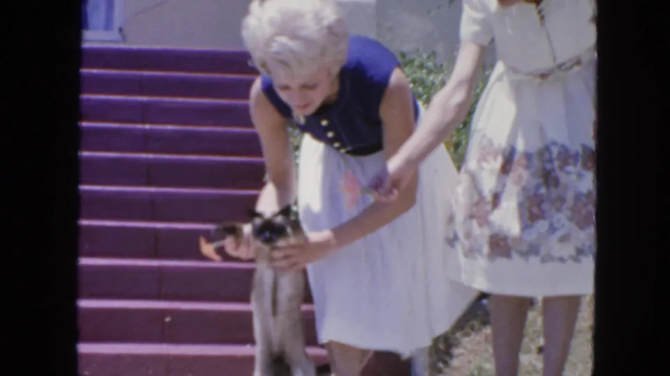 1963 San Francisco home-movie frame of two women beside porch steps as one steadies a small cat in bright sunlight.
