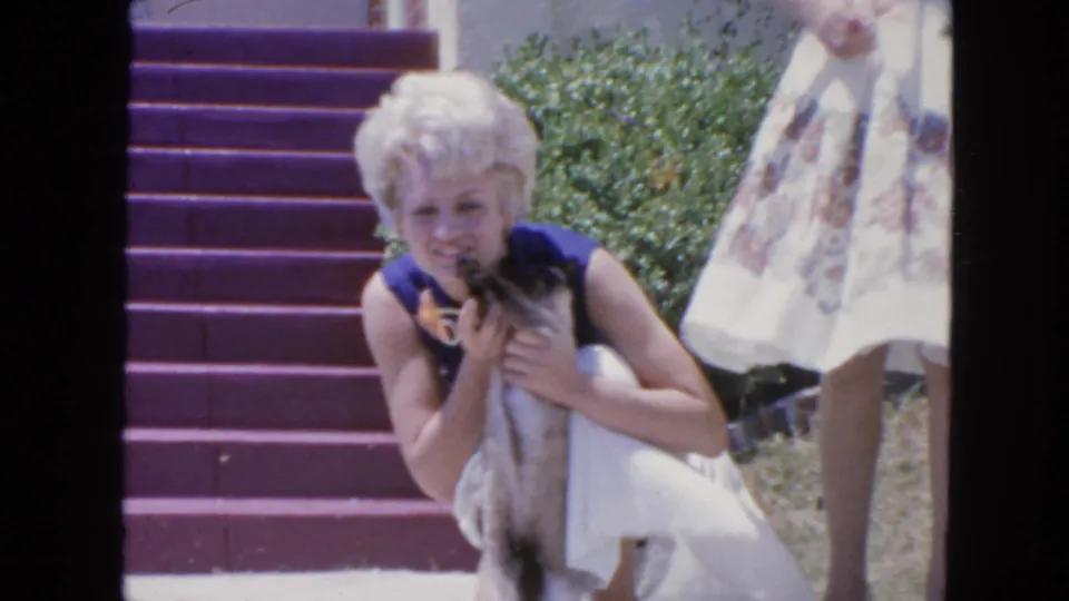 1963 San Francisco frame showing a woman smiling toward the camera while holding a small cat beside porch steps.