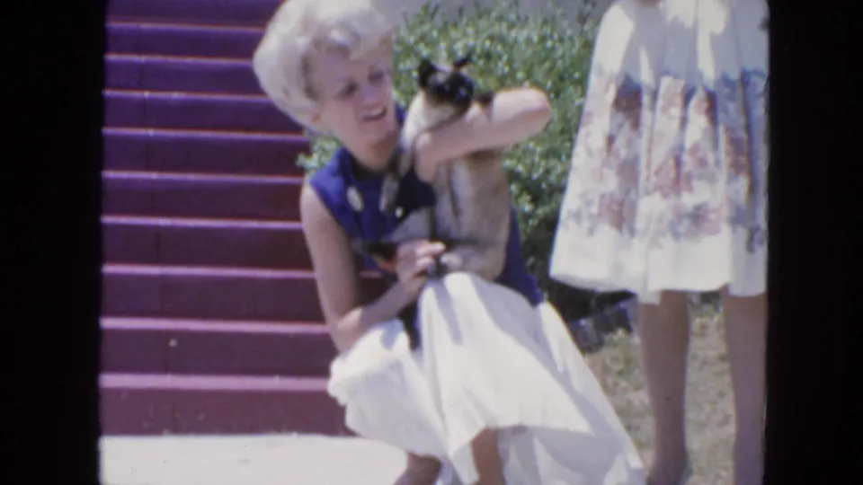 1963 San Francisco porch footage with a smiling blonde woman holding a Siamese cat near purple steps and a standing woman nearby.