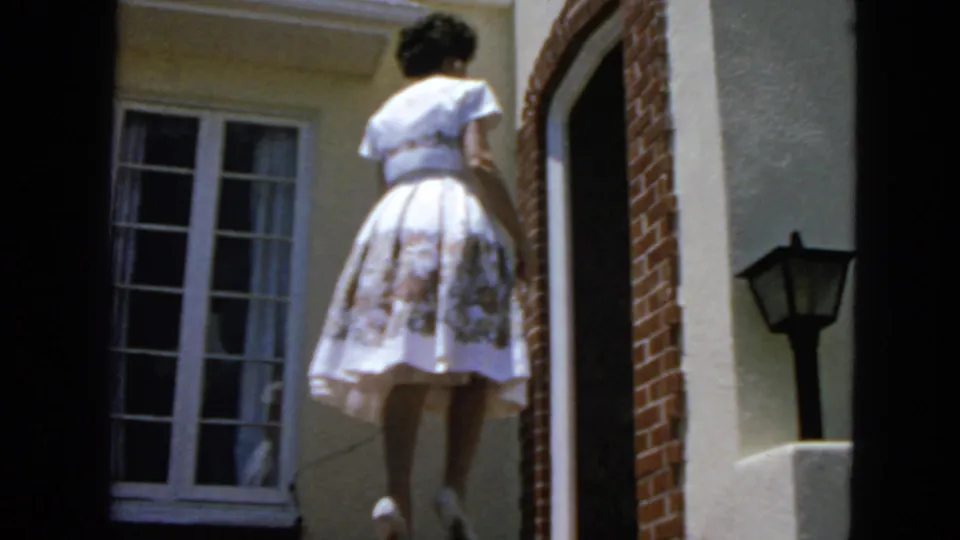 1963 San Francisco home-movie frame showing a woman in a white floral dress walking toward a brick doorway beside a window and wall lamp.