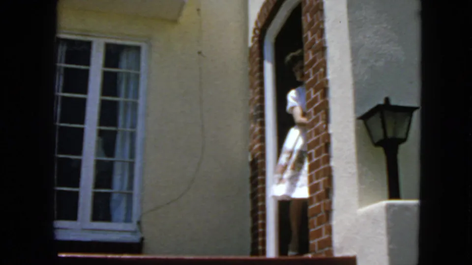 A woman in a light dress stands at the front entry of a San Francisco house, seen through dark window divisions in 1963 footage.