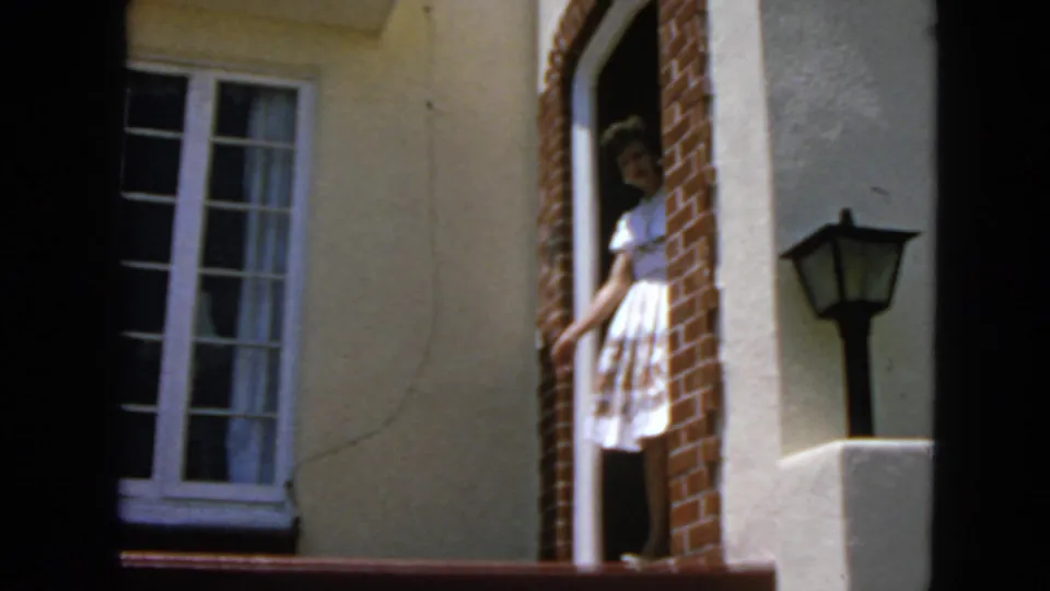 The woman is visible at the doorway of the stucco house, with brick edging, a window, and a porch lamp in the frame.