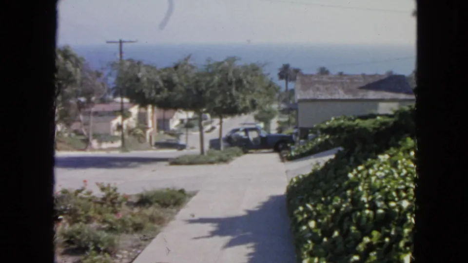 1963 8mm view of a San Francisco residential street from inside, with landscaping, low houses, parked car, and coastal horizon.