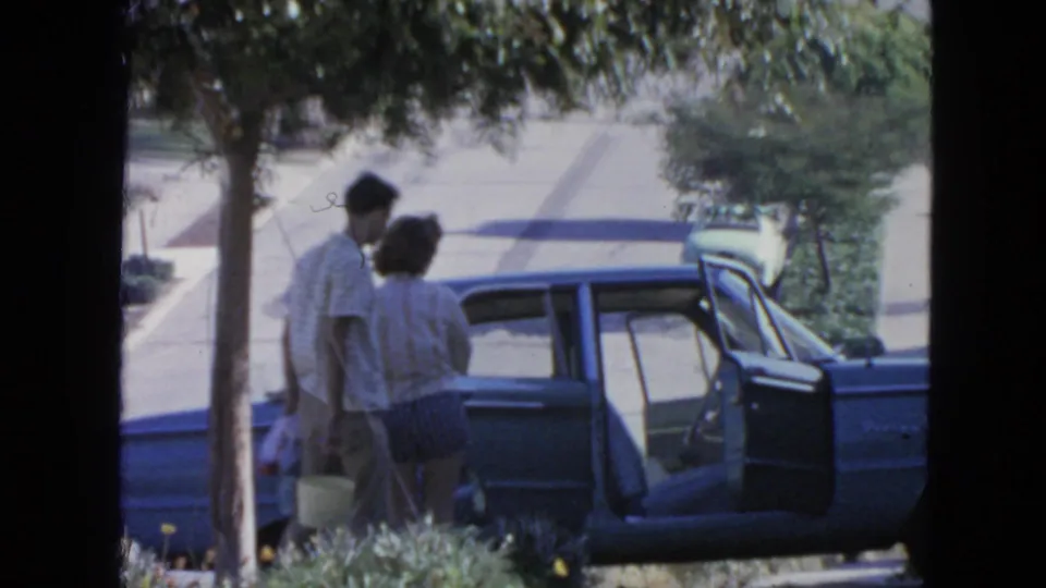 Two adults in 1960s casual clothing stand beside an open blue sedan in a San Francisco residential street scene.