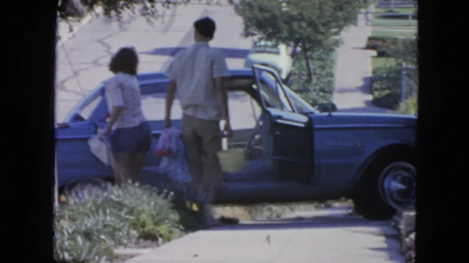 Two adults approach an open blue sedan in a 1963 San Francisco home-movie neighborhood scene.