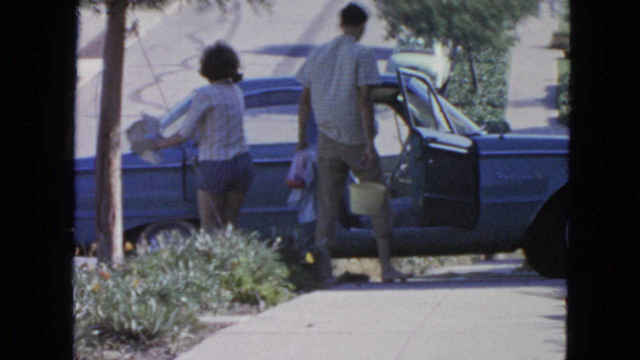 Two adults carry items beside a light blue sedan with an open door on a sunny street.