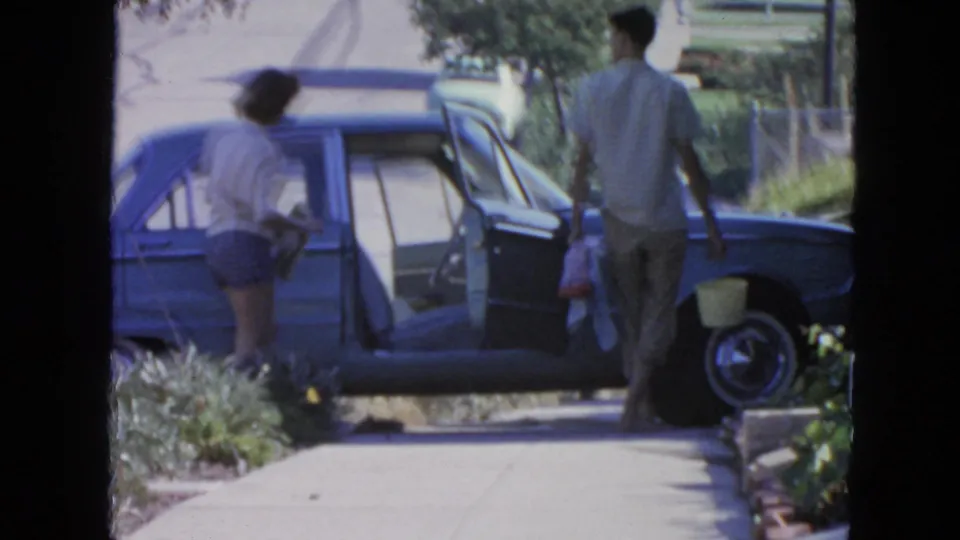 The woman stands near the open door of the blue sedan as the man moves with bags and a bucket on a sunny San Francisco street.