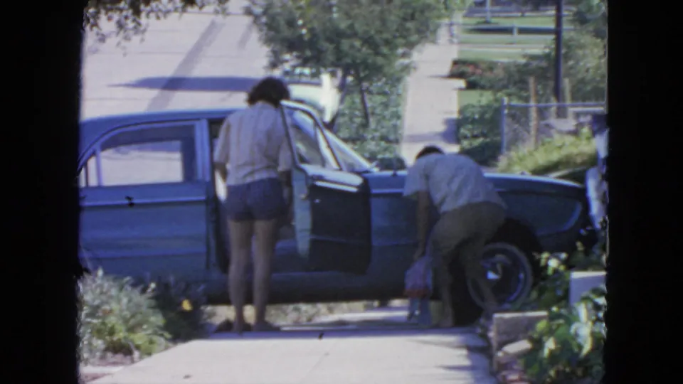 A woman waits beside the open blue sedan while a man bends down to gather or place bags beside the sidewalk.