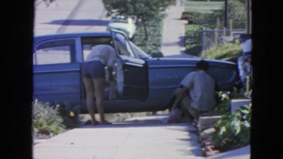 The woman leans into the blue sedan as the man crouches by bags along the sidewalk in this 1963 San Francisco home movie.