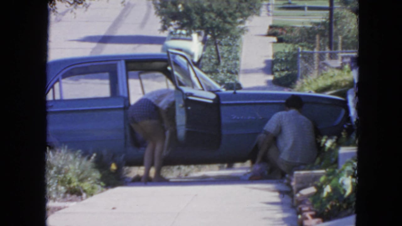 1963 San Francisco home-movie frame with a woman checking a blue sedan as a man crouches near the wheel on a residential sidewalk.