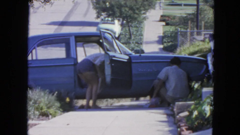 1963 San Francisco home-movie frame with a woman checking a blue sedan as a man crouches near the wheel on a residential sidewalk.