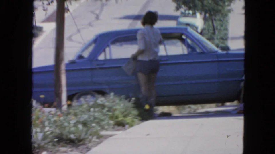 1963 San Francisco home-movie frame with a woman standing beside a blue sedan on a planted residential sidewalk.