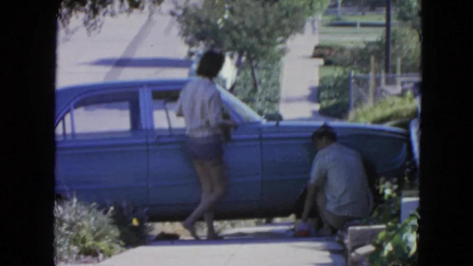 1963 San Francisco home-movie frame showing a woman beside a blue sedan and a man crouched near the wheel on a residential sidewalk.