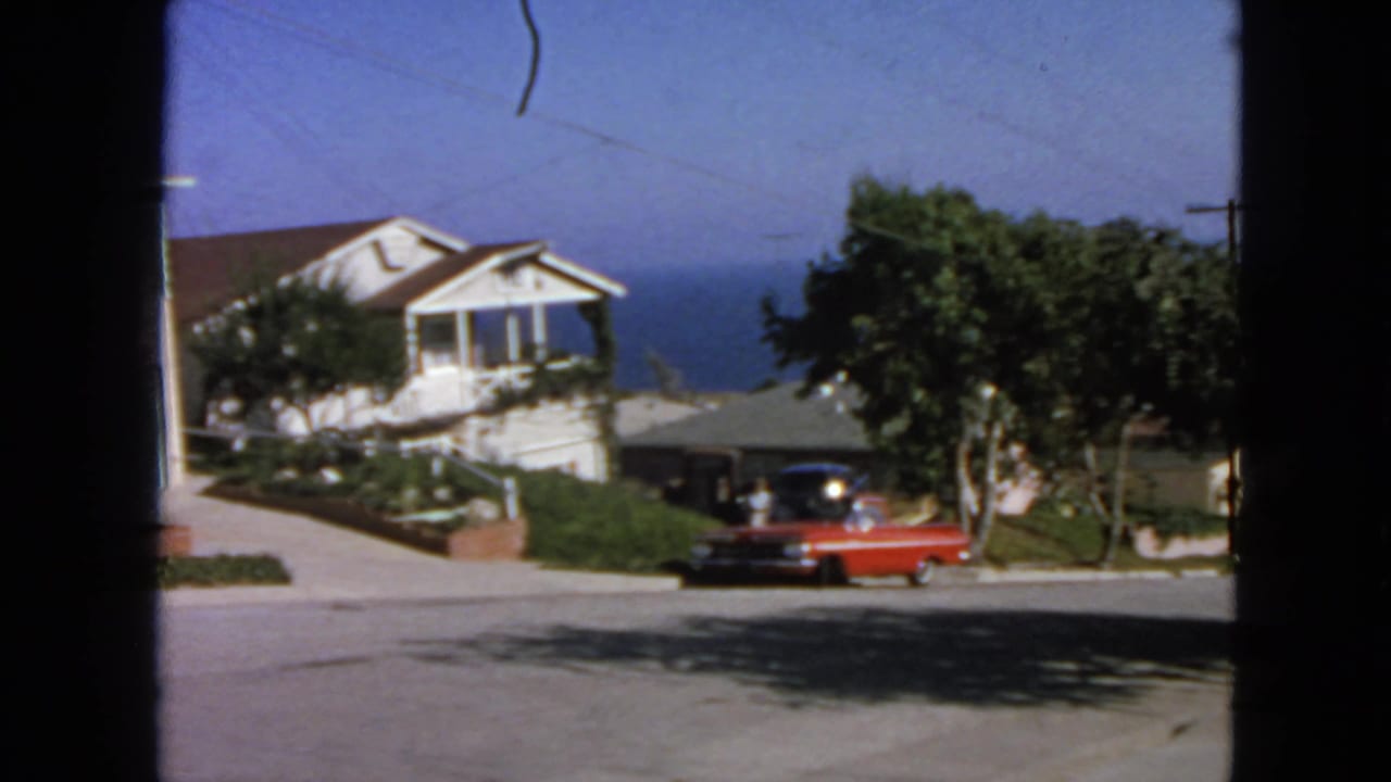 1963 San Francisco home-movie frame showing a red sedan near a white house, trees, a sloped street, and water in the distance.