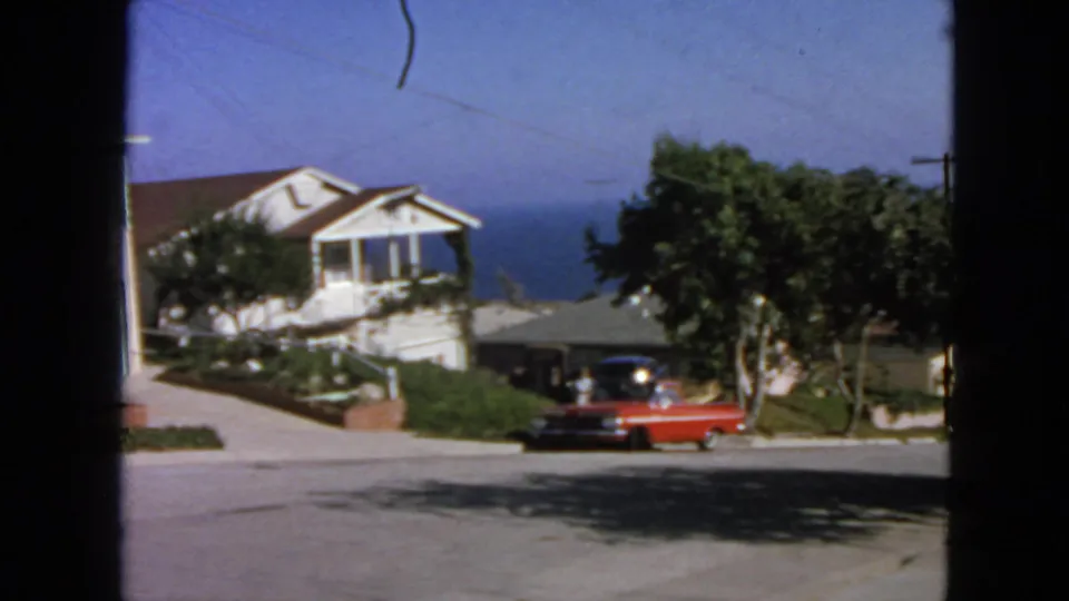 1963 San Francisco home-movie frame showing a red sedan near a white house, trees, a sloped street, and water in the distance.
