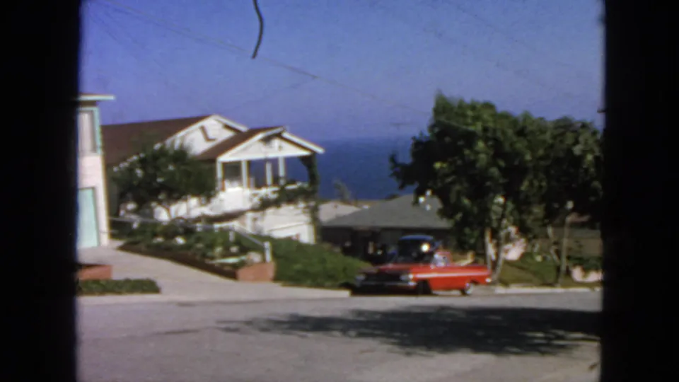 1963 San Francisco frame with an adult near a red sedan, residential houses, trees, and distant blue water behind the street.