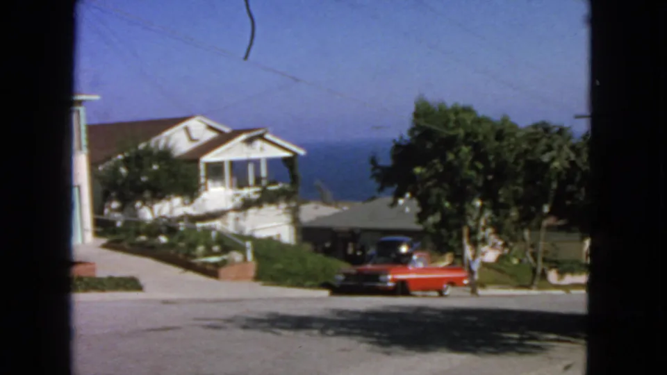 1963 San Francisco home-movie still of a red sedan by a curb as an adult figure stands nearby on a coastal residential block.