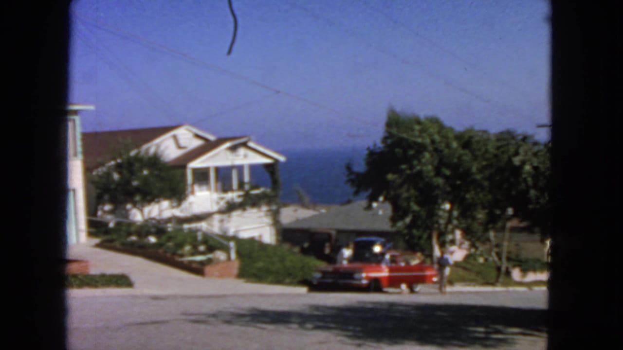 Residential street seen through a dark window frame with a red parked car, houses, trees, and ocean in the distance.