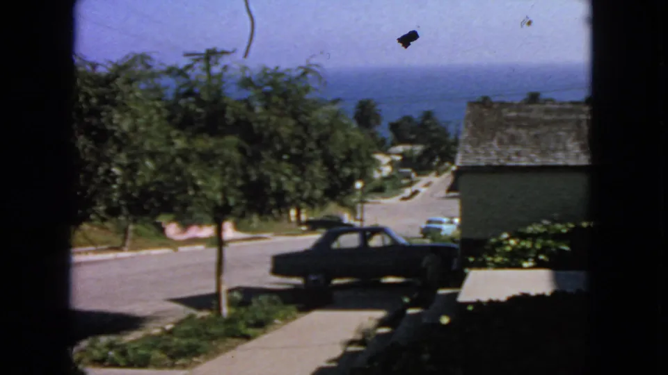 1963 San Francisco residential street with a dark sedan, leafy trees, sidewalk, nearby houses, and the ocean visible in the background.