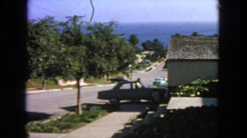 1963 San Francisco home-movie frame showing a dark sedan on a quiet residential street, with houses, trees, and ocean beyond.