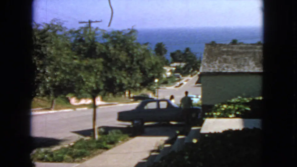 1963 San Francisco street view with a person near a dark classic sedan, houses, trees, sidewalk, and distant ocean.