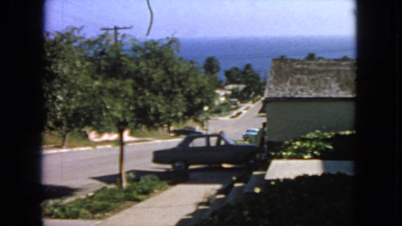 Residential street seen through a window with a dark sedan parked near a driveway and ocean beyond.