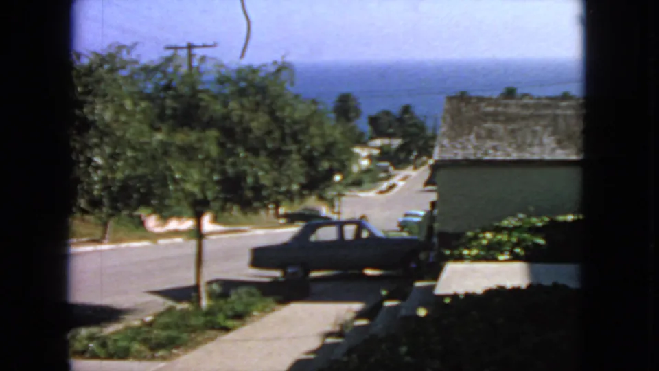 1963 San Francisco home-movie frame looking through a window at a dark sedan, street trees, neighboring houses, and the ocean horizon.