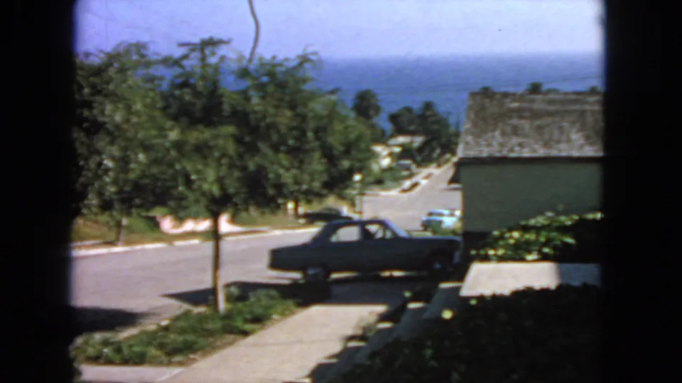 1963 San Francisco residential view through a window, showing a parked sedan, leafy street, sloping driveway, houses, and blue ocean beyond.