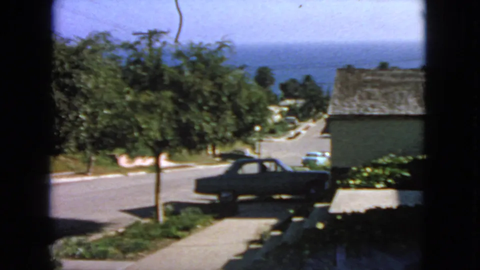 1963 San Francisco home-movie still of a dark car near a driveway, with street trees, a neighboring house, and distant ocean under bright daylight.