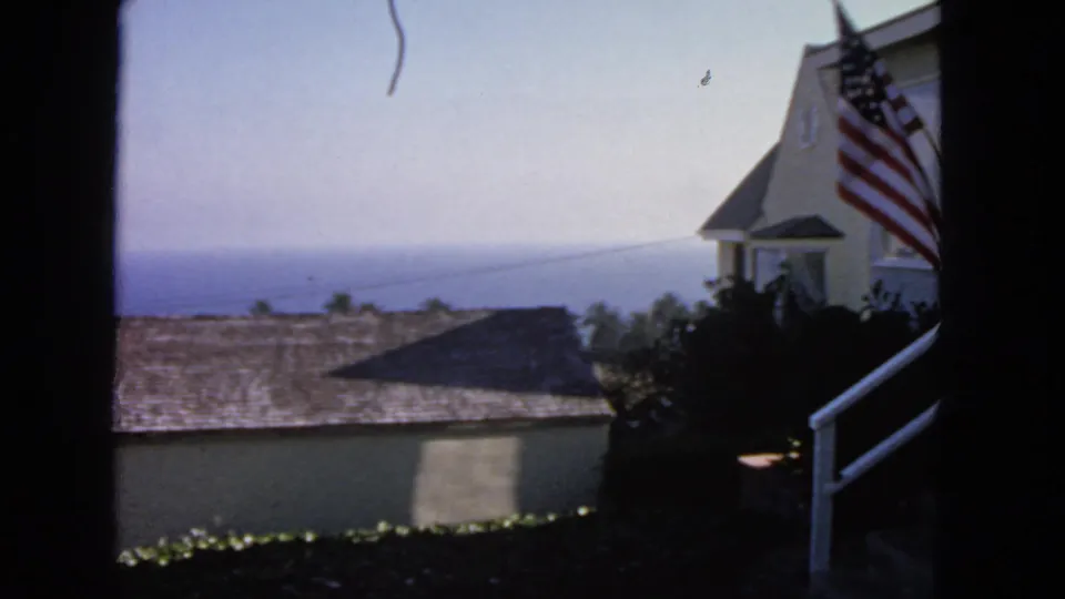 1963 San Francisco home-movie frame showing beige buildings, a porch flag, shingled roofs, and the ocean in the distance.