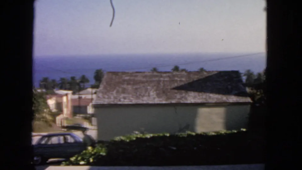 1963 San Francisco frame showing a dark sedan passing below beige buildings, with palm trees and ocean beyond.