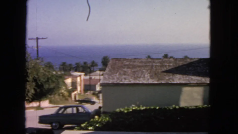 1963 San Francisco home-movie frame with a dark sedan, beige buildings, utility pole, palm trees, and ocean horizon.
