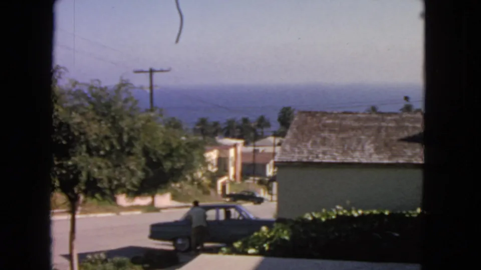 1963 San Francisco frame with a man beside a curbside sedan, seen from indoors toward houses, palms, utility lines, and the distant Pacific Ocean.