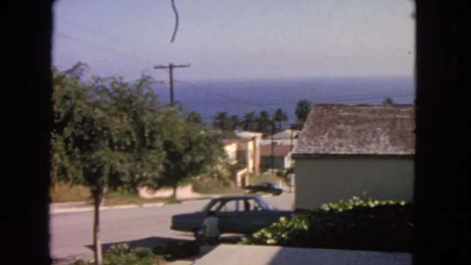 A 1963 San Francisco home-movie view of a man near a parked sedan on a residential street, framed by a window with coastal water in the background.