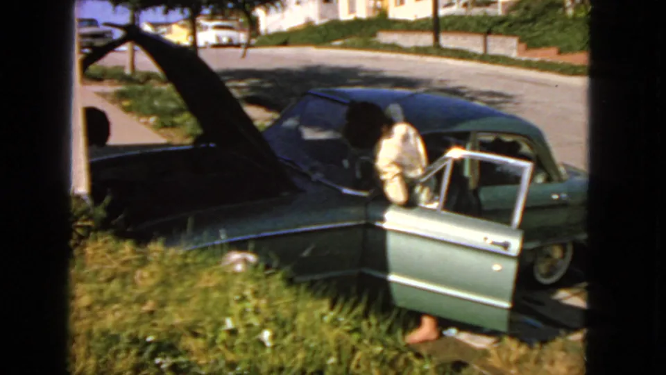 1963 San Francisco frame showing a man leaning at an open sedan door while the hood is raised, with sidewalk grass and residential street details visible.