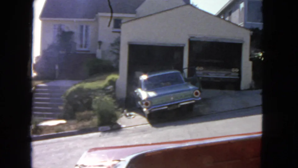 Sunlit 1963 San Francisco street frame with a blue car, sloped driveway, shrubs, and open residential garage.