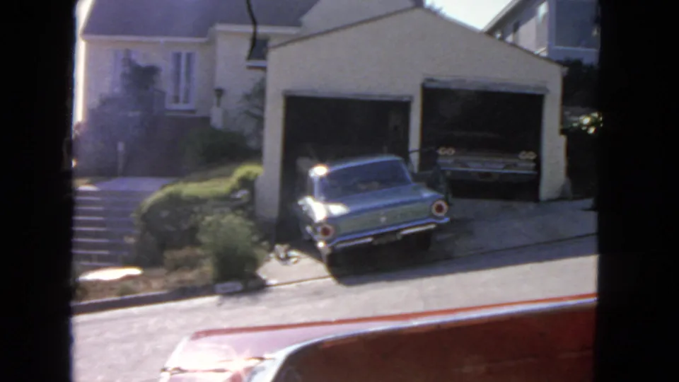 1963 San Francisco home-movie still of a blue car by a residential garage, with a red vehicle edge crossing the foreground.