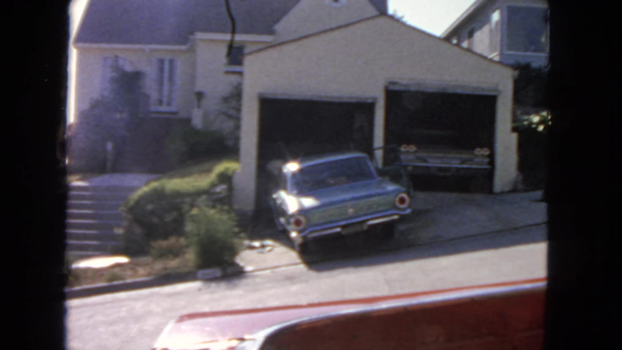 Light blue classic car parked in a driveway beside a two-car garage and house.