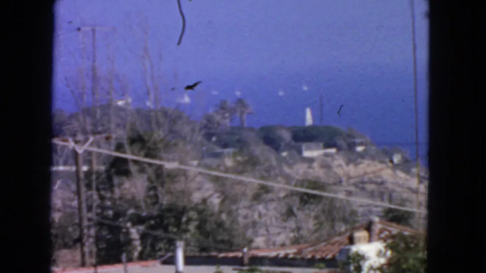 1963 San Francisco coastal frame with hillside vegetation, foreground rooflines, ocean, and a distant white tower or lighthouse.