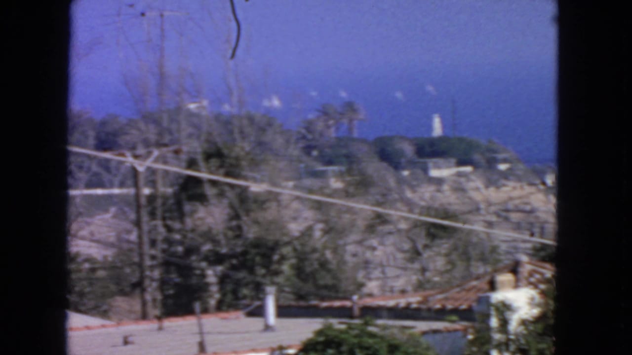 Coastal hillside with utility lines, blue sky, and a distant white tower above a garden wall.