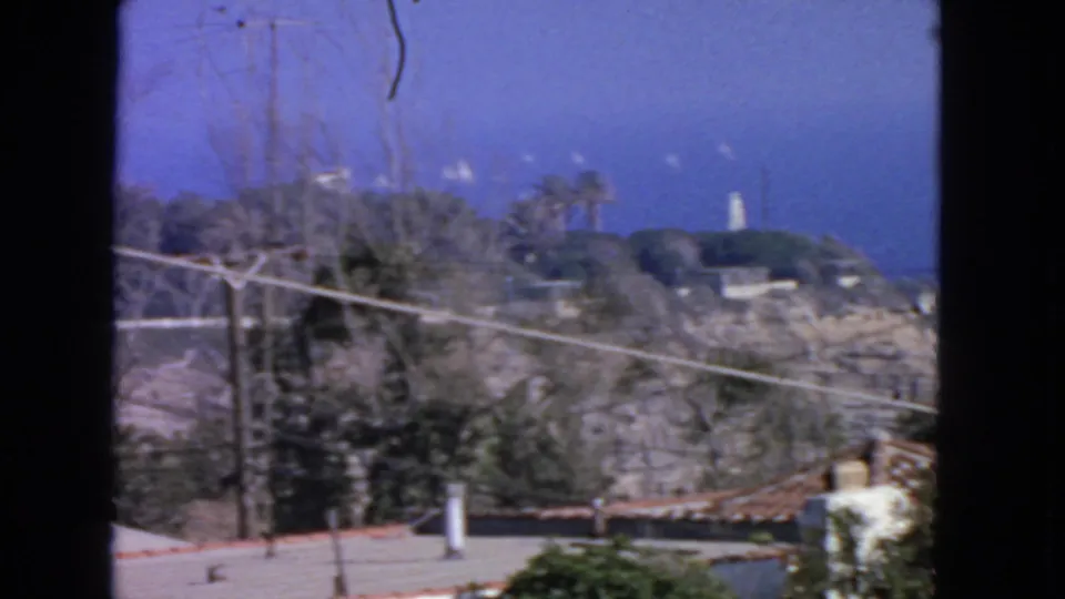 1963 8mm San Francisco coastal view showing hillside brush, utility lines, blue sky, and a distant white tower beyond a low wall.